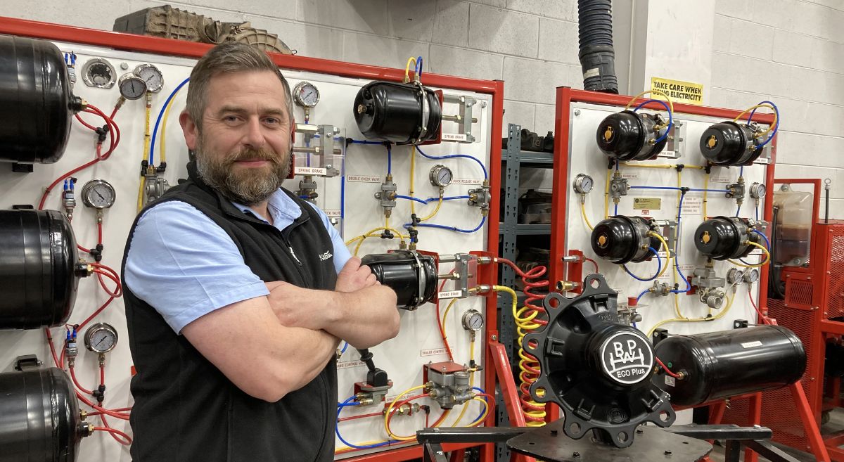 A man with a beard, wearing blue short sleeved shirt and black gilet, arms folded, smiling to camera with motor engineering articles in background